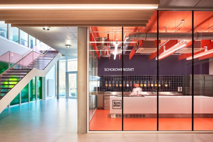 View of a modern, brightly lit chocolate workshop with a person working at a counter and large glass windows displaying "Schoko Werkstatt"