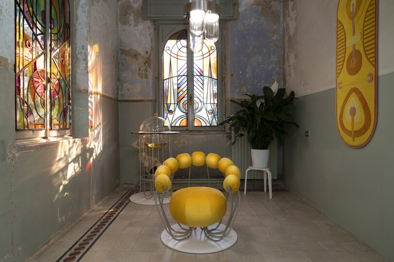 A room with colorful stained glass windows, a yellow chair, a plant on a white stool, and decorative wall art.