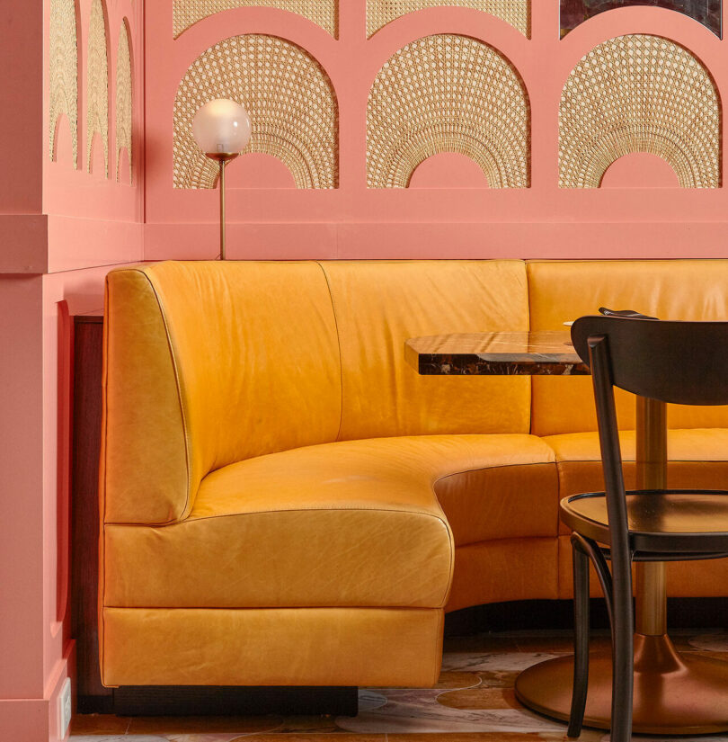 A cozy seating area features an L-shaped yellow couch with a round wooden table and a black chair, set against a pink wall with decorative arches and a globe lamp.