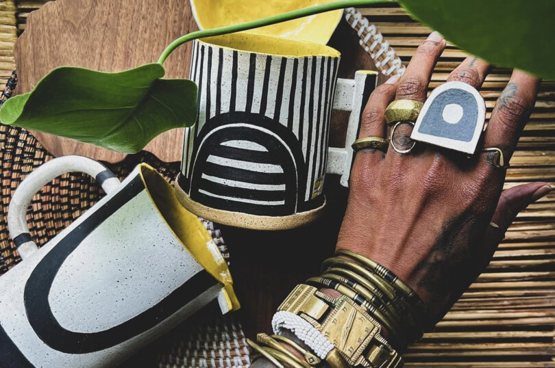 A hand with rings and bracelets rests on a woven surface. Nearby are two ceramic mugs with black and white geometric patterns and yellow interiors. A leaf partially covers one mug.