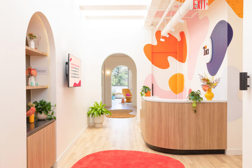 Bright lobby with modern decor, featuring colorful wall art, a wooden reception desk, plants, and an arched hallway leading to a sunlit room.