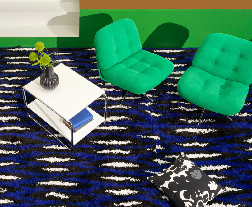 Two green chairs and a white table on a black and blue patterned rug, paired with stylish home furnishings like a black vase filled with green flowers. A chic black and white floral cushion rests elegantly on the rug.