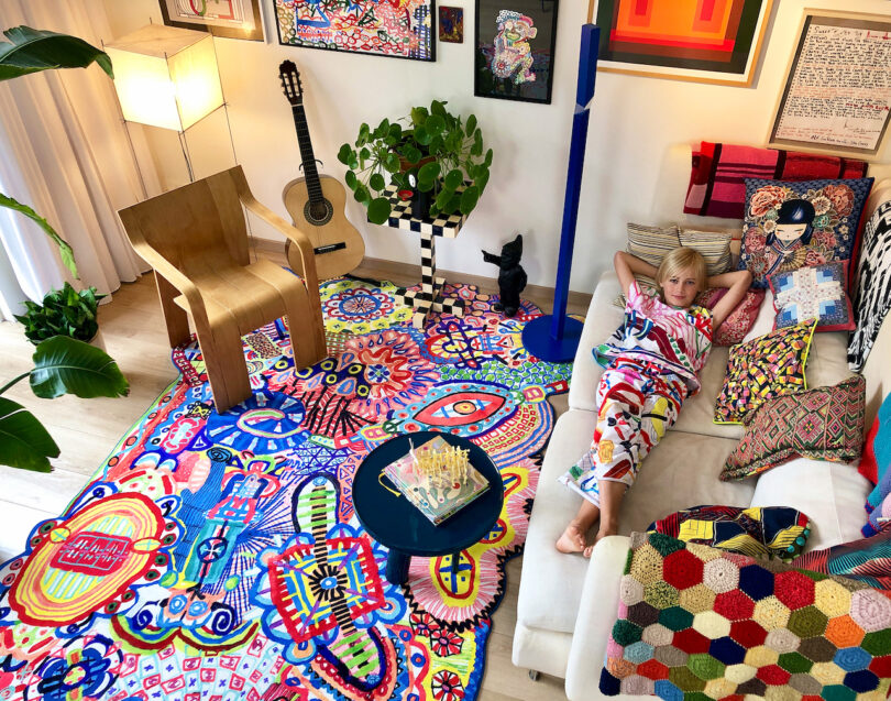 A person in colorful attire relaxes on a white couch, surrounded by art, plants, and a guitar, blending seamlessly with the vibrant home furnishings that complete the lively ambiance of the room.