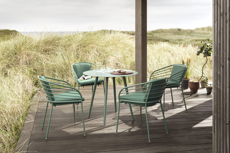 Four green chairs surround a small round table on a wooden deck overlooking grassy dunes and a cloudy sky.