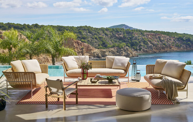 Outdoor patio with wicker furniture, a coffee table, and cushions on a striped rug overlooking a pool and scenic ocean view with hills in the background.