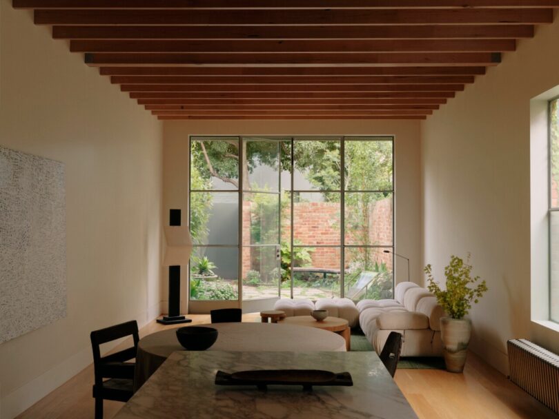 A modern living room with a marble table, black chairs, and a beige sofa. Large windows overlook a garden with trees and a brick wall. Exposed wooden beams line the ceiling