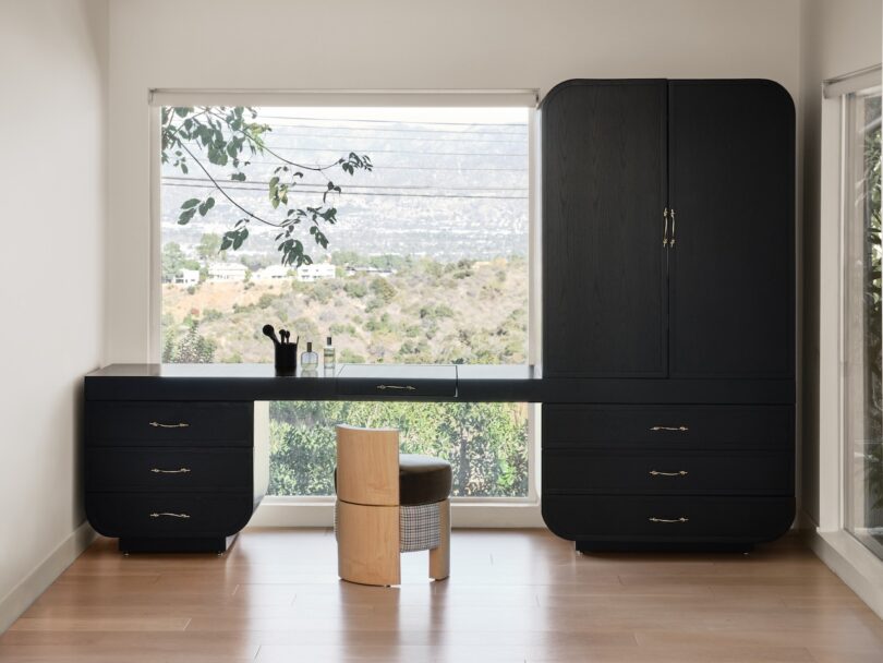 A minimalist room features a long black desk with drawers, a wooden chair, and a large window showcasing a mountain view