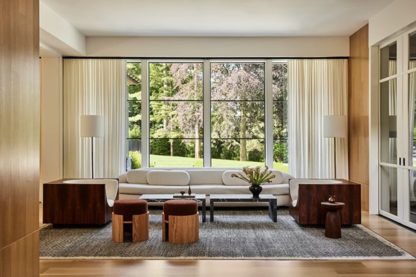 Modern living room with large windows, a white sofa, wooden side tables, small brown stools, and a central coffee table on a gray rug. Greenery is visible outside the windows