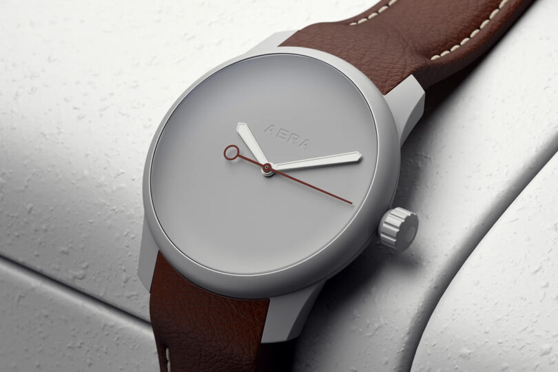 Minimalist wristwatch with a brown leather strap and a simple gray dial, featuring two hands and no indices, placed on a textured surface.