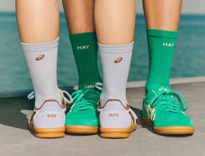 Three people wearing green and gray HAY sneakers with matching ASICS and HAY socks stand on a boardwalk near a body of water.