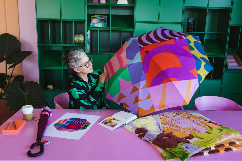 A person with gray hair and glasses examines a colorful umbrella at a pink table. The background features green shelves with various items.