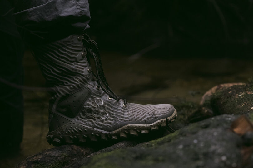 A person wearing rugged, textured boots stands on moss-covered rocks beside a stream.