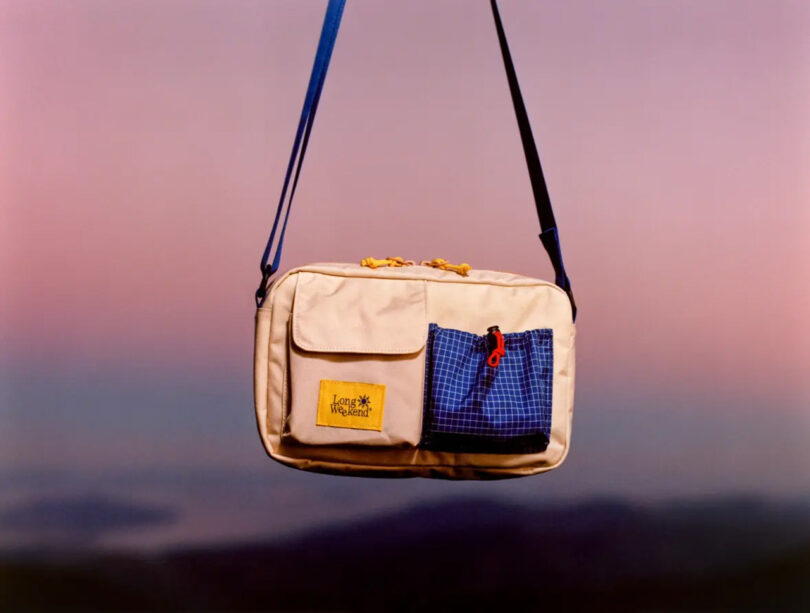 A beige and blue crossbody bag with front pockets and a "Have a Good Weekend" patch, set against a blurred pink and purple background.