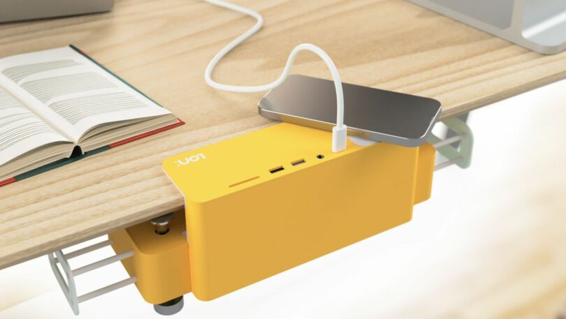 Une station de recharge jaune fixée à un bureau avec un smartphone connecté via un câble. Un livre ouvert se trouve à proximité.