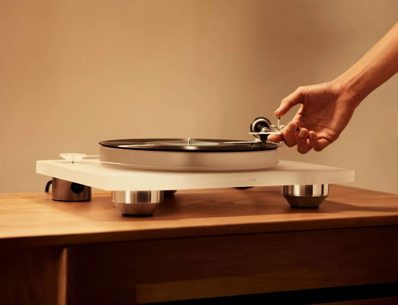 Une main ajuste le bras d'un plateau tournant blanc avec un disque vinyle noir sur une surface en bois.