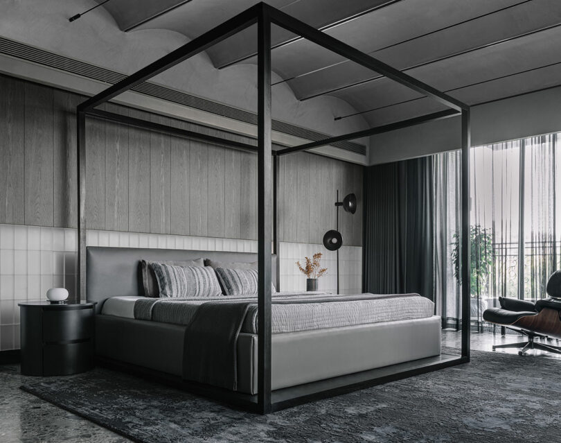 Modern bedroom with a four-poster bed, neutral bedding, a round bedside table, wall-mounted lights, and a large window with sheer curtains.