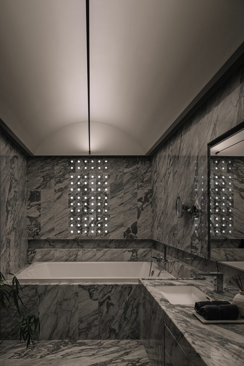 Luxurious marble bathroom with a large bathtub, a sink, and a well-lit mirror. The walls and floor are covered in grey marble tiles. A window grid provides ambient light.