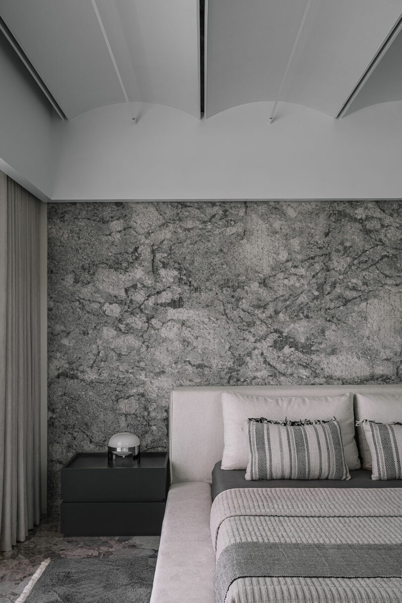 Modern bedroom with a gray marble accent wall, light gray upholstered bed, striped bedding, and a sleek black nightstand with a round lamp.