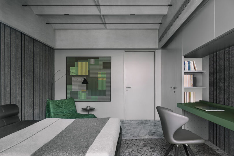 A modern bedroom with a minimalist design, featuring a bed, gray chair at a small green desk, abstract wall art, and a green armchair against textured walls and flooring.