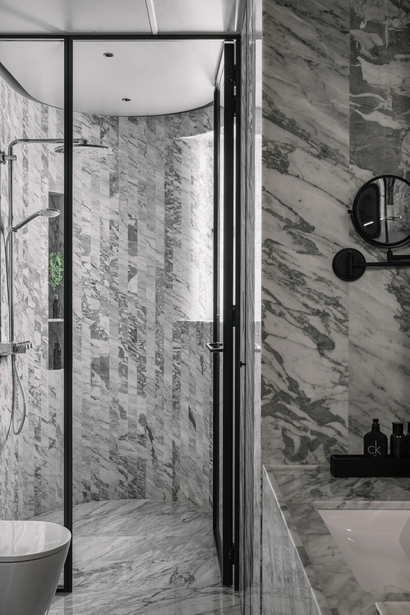 Luxurious bathroom with grey marble walls and floors, a glass shower enclosure, a white bathtub, and a modern sink area with a round mirror.