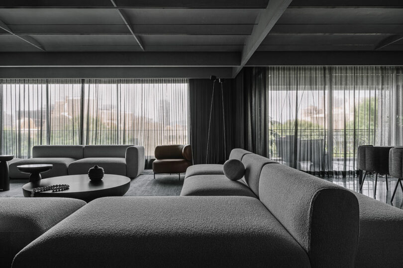 A spacious, modern living room with gray sofas, a round coffee table, and large windows with sheer curtains, offering a view of greenery outside.