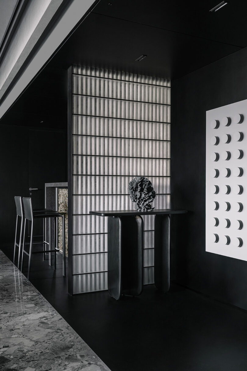 Modern interior with a sculpted bust on a black console table, flanked by a textured wall. High chairs and a patterned art piece complete the space.