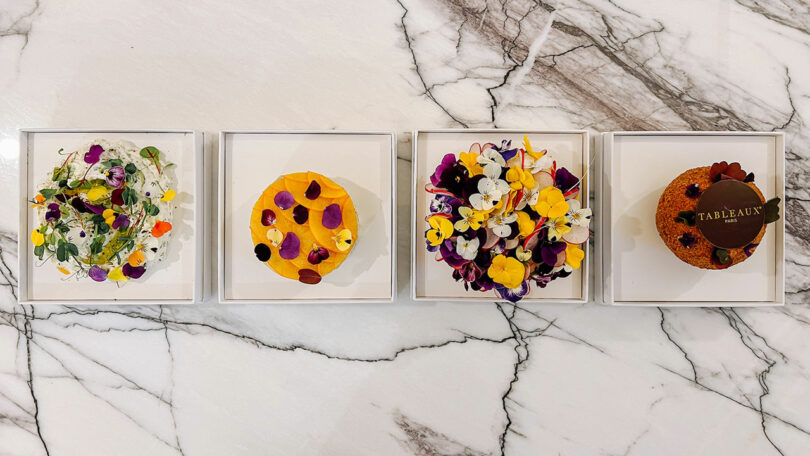 Four floral-topped desserts displayed in square white boxes on a marble surface.