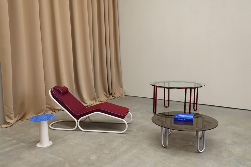 A modern lounge chair, a Yonder Table, and a small blue stool are arranged on a gray floor with beige curtains in the background.