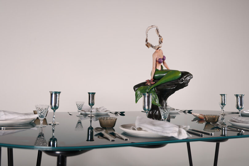 A sculpture of a headless mermaid on a rock graces the Tubulaa glass dining table, elegantly set with cutlery, glasses, and napkins.