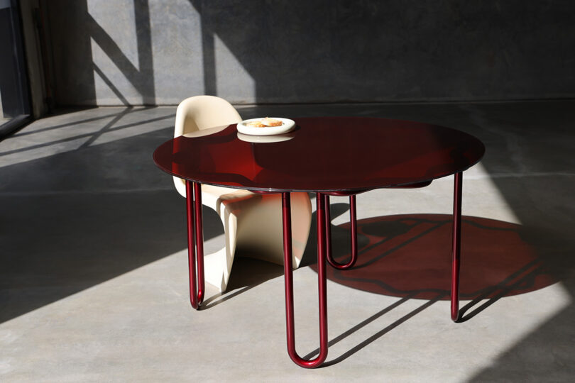 The Yonder Table captures modern elegance with its red oval design and sleek curved legs, accompanied by a minimalist white Tubulaa chair in a sunlit room. A small bowl graces the table's surface, completing the chic setting.