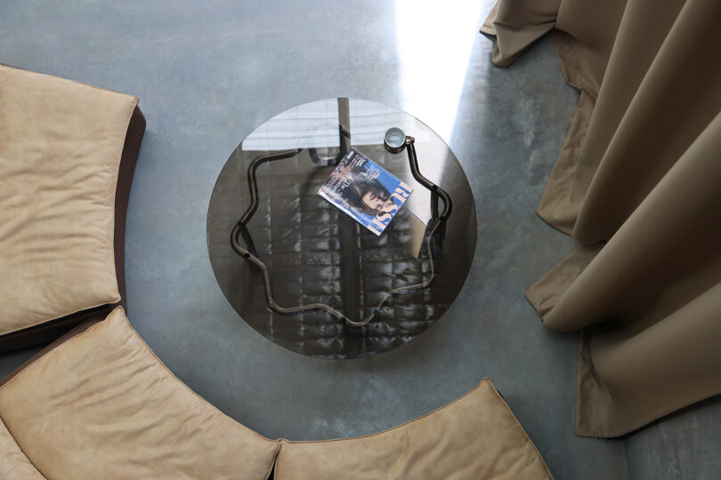 A round glass Yonder Table with a magazine and magnifying glass on top sits gracefully between beige curved sofas on a polished gray floor, complemented by beige curtains.