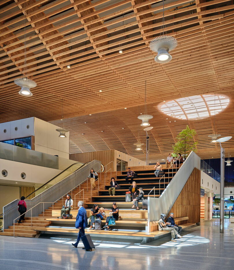 Des personnes assises sur un grand escalier intérieur en bois avec un plafond ouvert et une lucarne moderne, qui rappelle le style architectural de ZGF. Une personne passe avec des bagages.