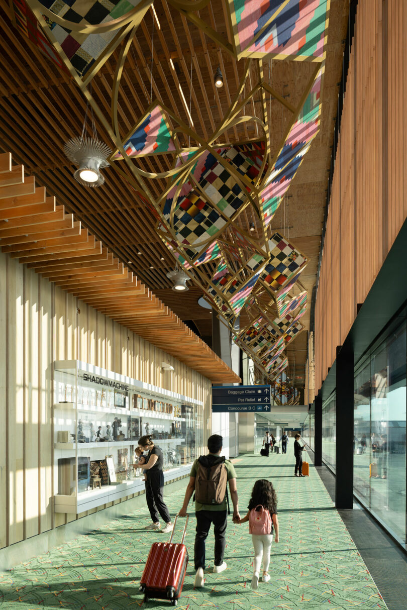 Un homme et un enfant se tenant la main traversent un terminal d'aéroport conçu par ZGF, présentant un décor géométrique coloré au-dessus et une vitrine sur la gauche.