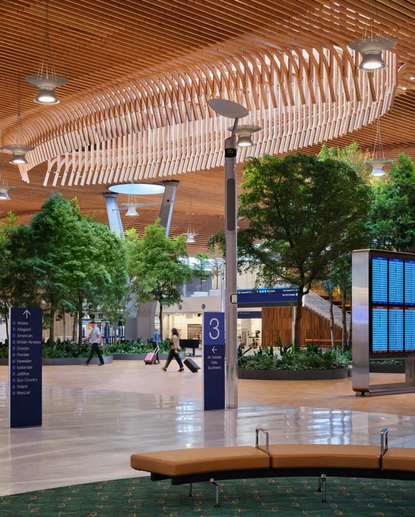 Un terminal d'aéroport moderne conçu par ZGF présente un superbe plafond en bois, des arbres intérieurs luxuriants, de nombreux sièges et un tableau de départ numérique élégant. Les passagers se promènent tranquillement dans l’espace aéré avec leurs bagages en remorque.