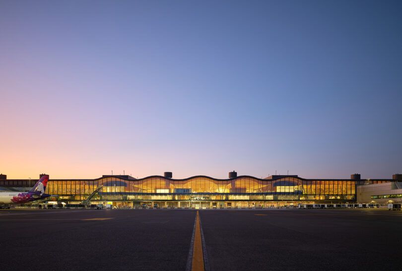 À la tombée du jour, la vue extérieure du terminal aéroportuaire conçu par ZGF prend vie, avec un avion garé sur la gauche et le bâtiment magnifiquement éclairé sous un ciel dégagé.