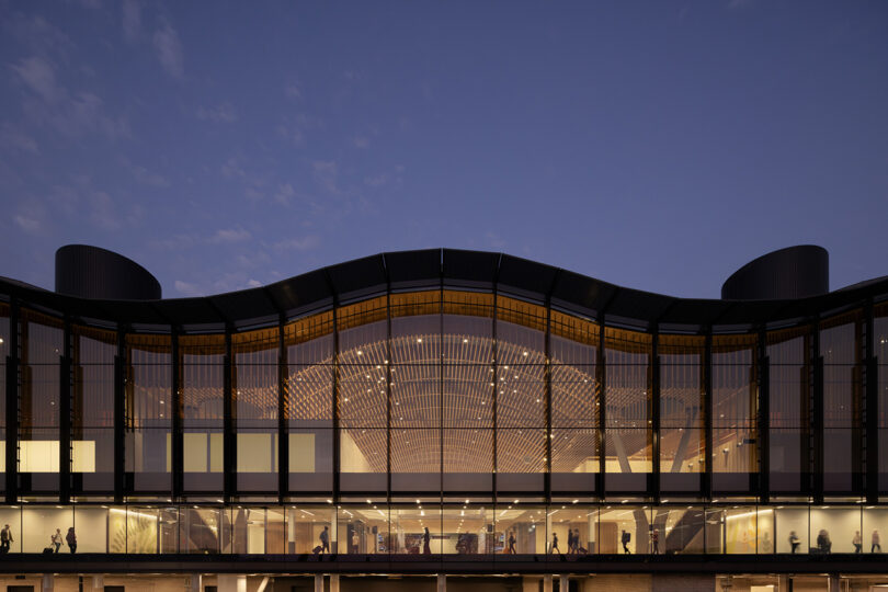 Vue nocturne d'un bâtiment moderne conçu par ZGF avec de grandes fenêtres en verre, mettant en valeur l'éclairage intérieur et les silhouettes de personnages à l'intérieur.