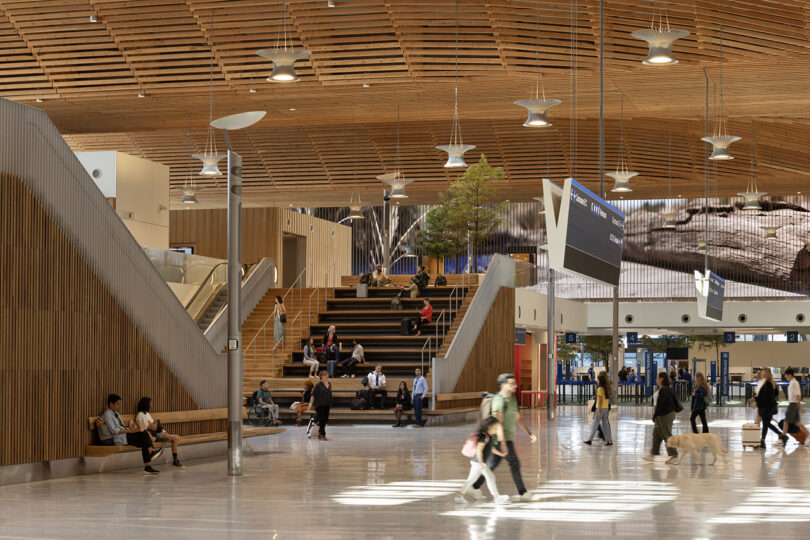 L'intérieur d'un grand terminal d'aéroport moderne conçu par ZGF présente un superbe plafond en bois. Les passagers avec bagages déambulent et se détendent sur les bancs et les marches, tandis que de grands écrans affichent efficacement les informations de vol.