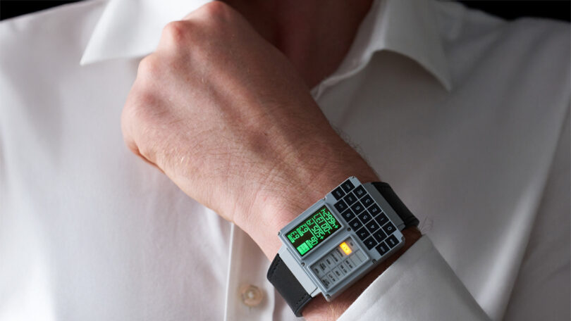 Person wearing a novelty watch designed like a miniature computer, featuring a digital screen and keypad, on their wrist over a white shirt.