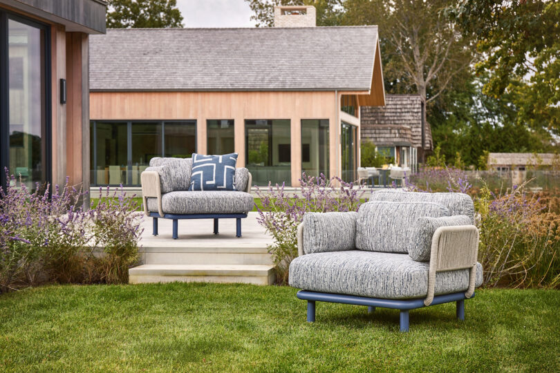 Two cushioned outdoor chairs on a lawn, facing a modern wooden house with large windows, surrounded by purple flowering plants