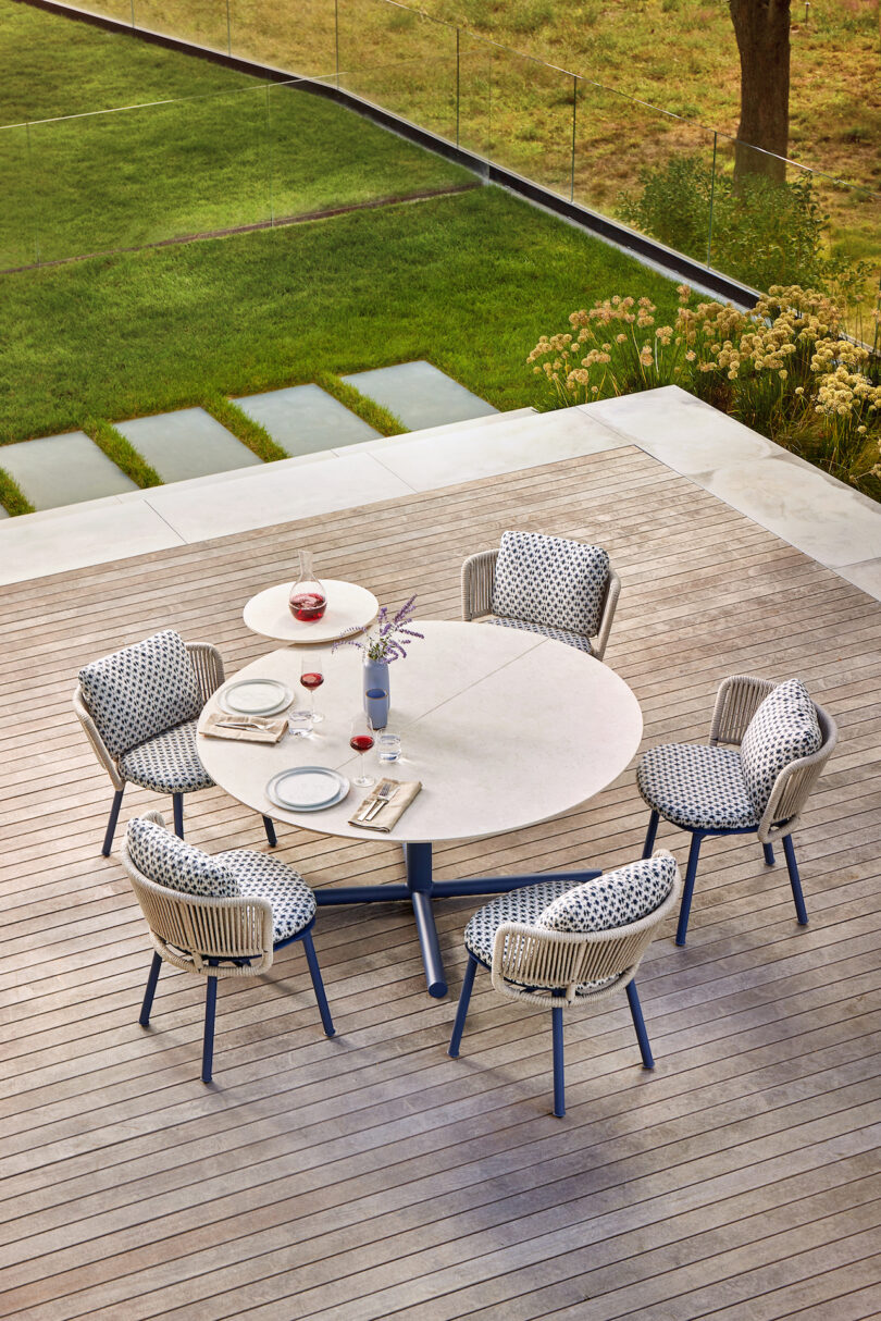 Outdoor patio with round table set for a meal, surrounded by five cushioned chairs on wooden decking, overlooking a grassy area