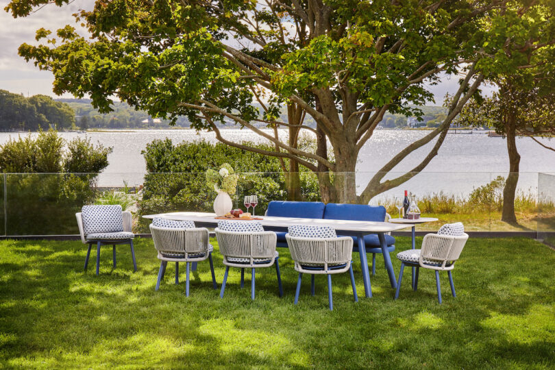 Outdoor dining setup with a table and eight chairs on a grassy area. There are trees and a lake in the background. Wine, glasses, and a vase with flowers are on the table