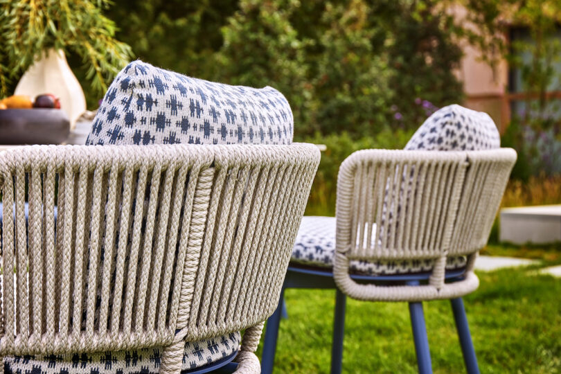 Two outdoor chairs with rope detailing and patterned cushions are set on a grassy area with trees in the background