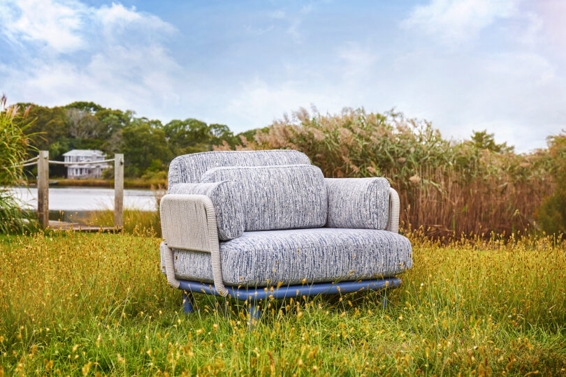 A blue and white sofa sits on green grass near a body of water, with trees and reeds in the background under a cloudy sky