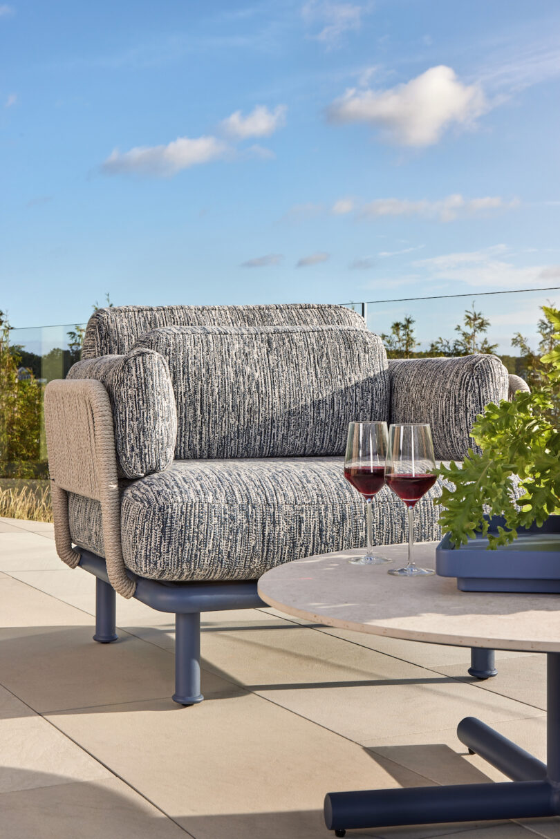 Outdoor patio with a cushioned chair, two glasses of red wine on a round table, and potted greenery, against a blue sky background