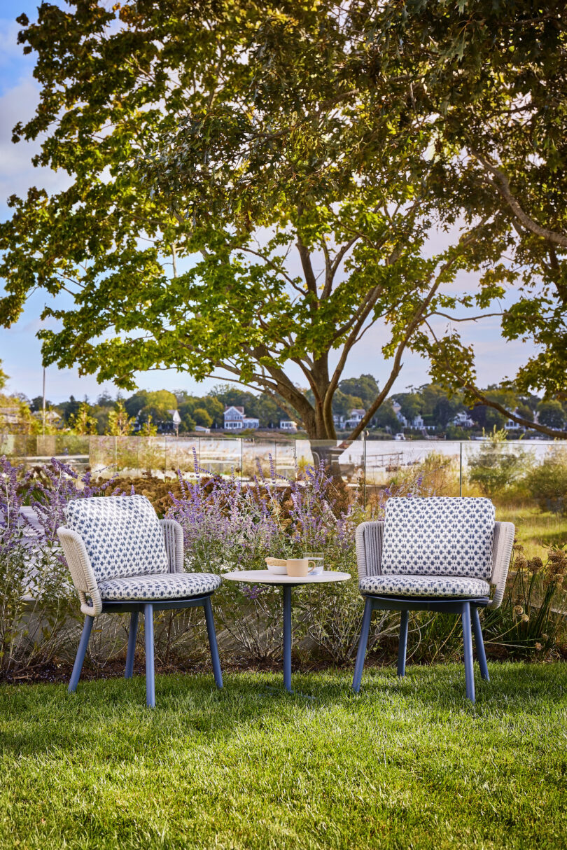 Two patterned chairs and a small round table on a grassy lawn under a large tree, with a scenic background of water and greenery