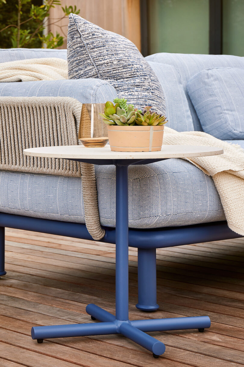 A blue outdoor sofa with a gray cushion and cream-colored blanket on a wooden deck. A small round side table holds a potted succulent and a clear glass