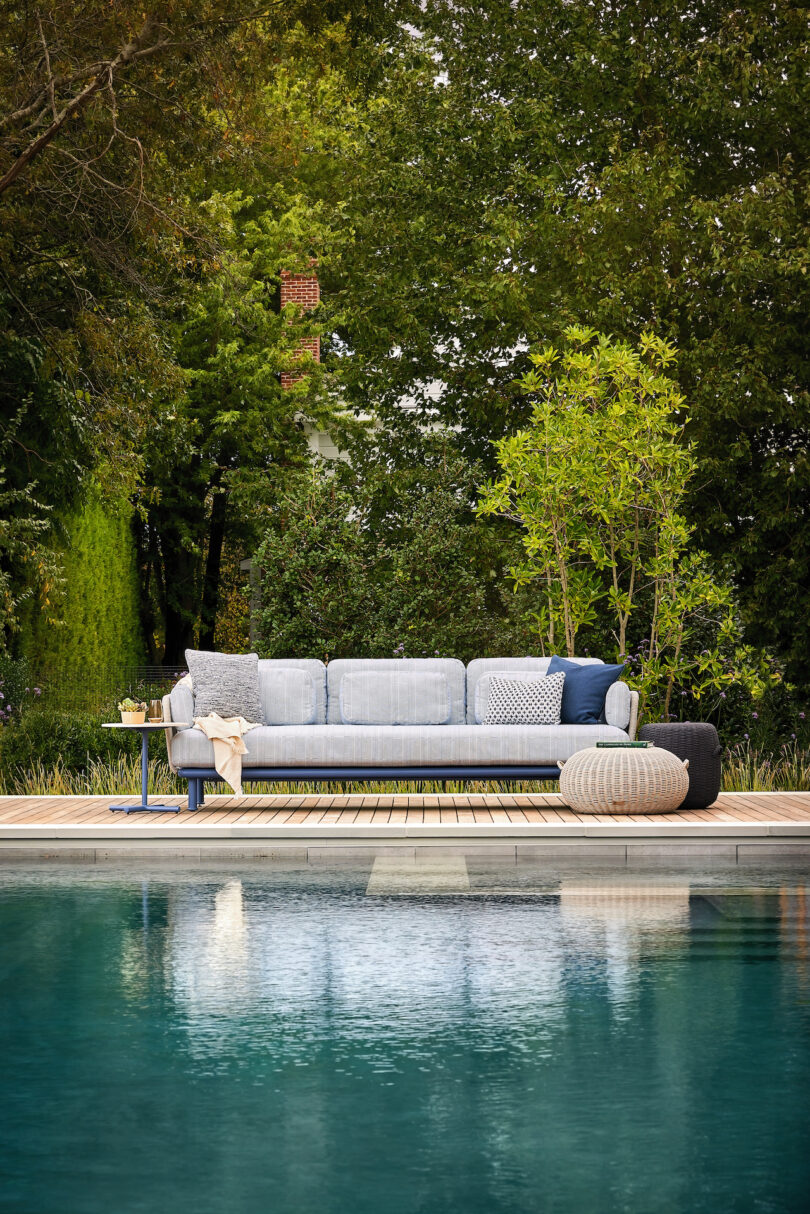 A modern outdoor sofa with cushions and a small side table, set on a wooden deck overlooking a calm pool, surrounded by lush greenery