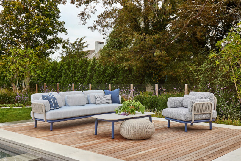 Outdoor patio with a sofa, chair, and coffee table on a wooden deck, surrounded by green trees and shrubs. A pouf and cushions are placed on the furniture