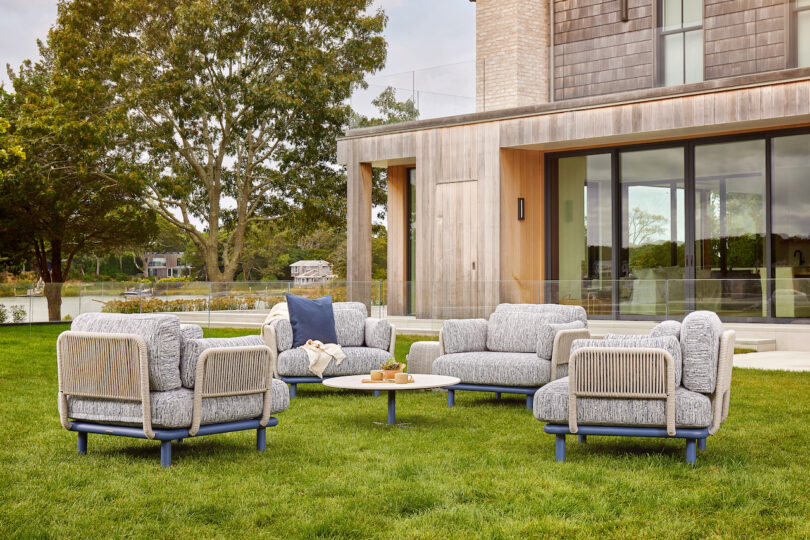 Outdoor seating area with four cushioned chairs around a circular table on a grassy lawn. A modern building with large windows is in the background