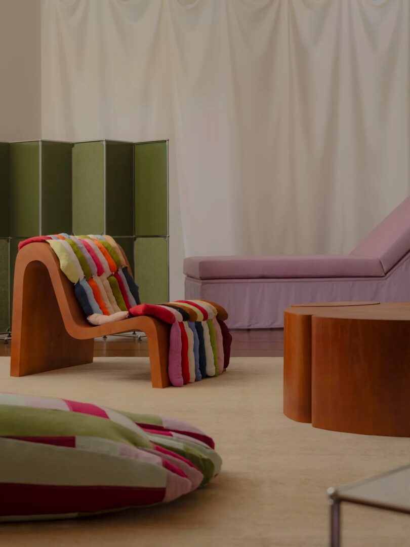 A modern room with a wood chair featuring colorful cushions, a lavender daybed, green room divider, and a beige rug with floor cushions